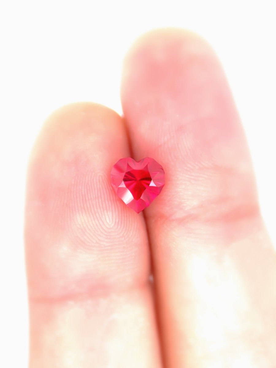Heart-shaped red sapphire held between two fingers on a white background for scale
