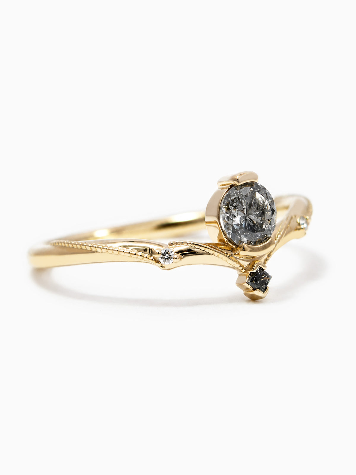 A gold ring designed like a branch, featuring a large round dark gray stone in the center and a small dark stone accent, set against a clean white background.