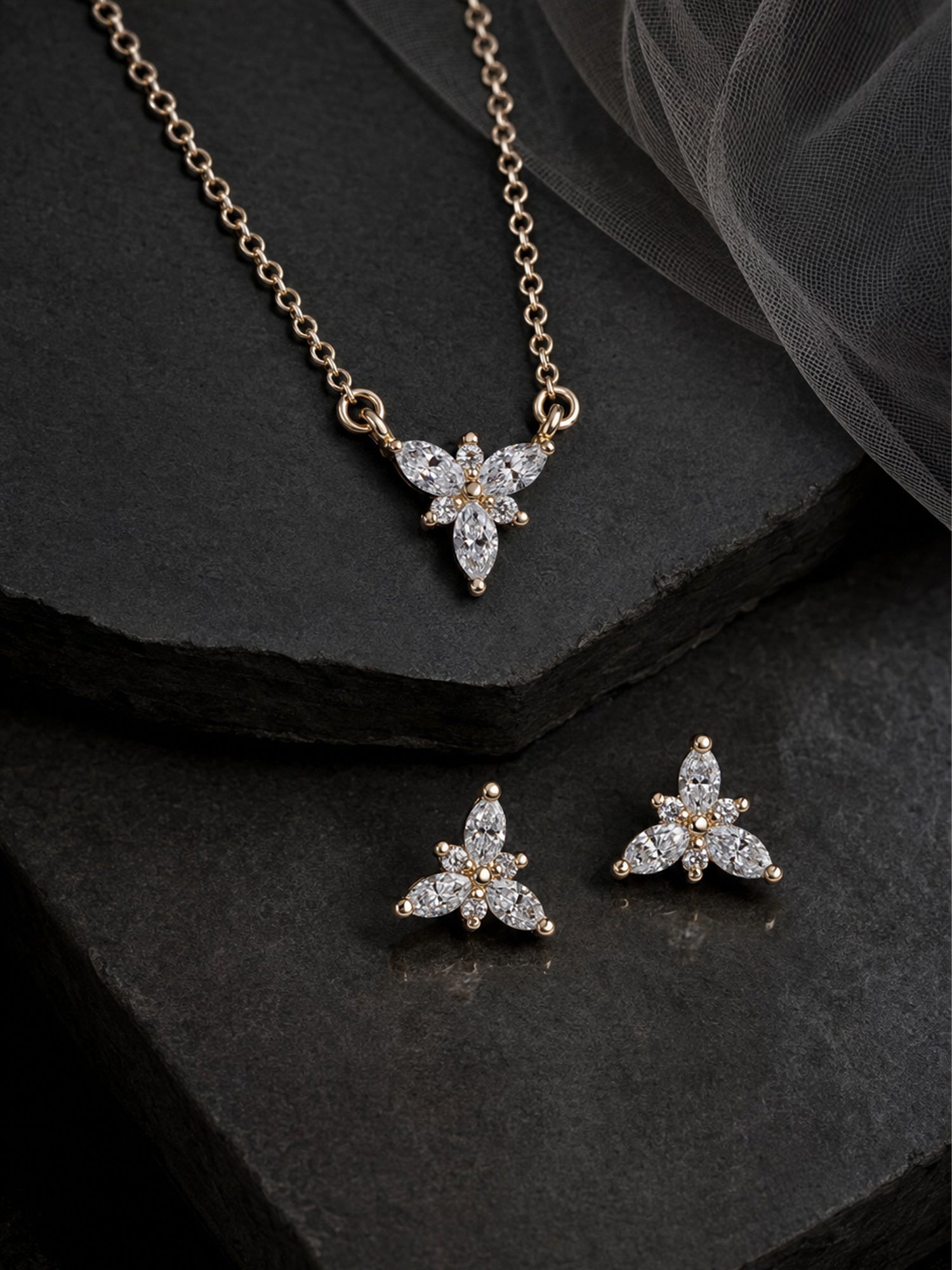 A gold-tone jewelry set featuring a pendant necklace and matching stud earrings, both with a butterfly or flower-like motif set with clear stones, displayed on dark textured stone slabs with sheer fabric in the background.