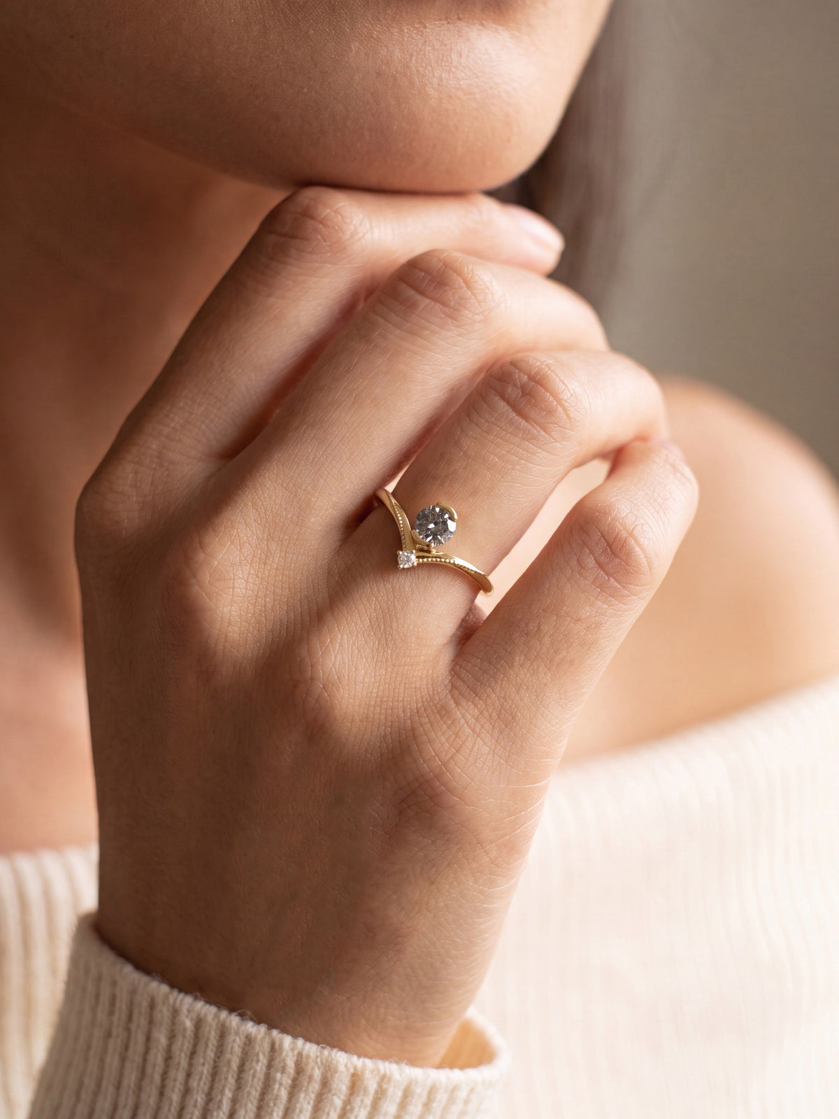 A close-up of a person's hand wearing a delicate gold ring with a round clear gemstone and a small accent stone, set against a soft neutral background.