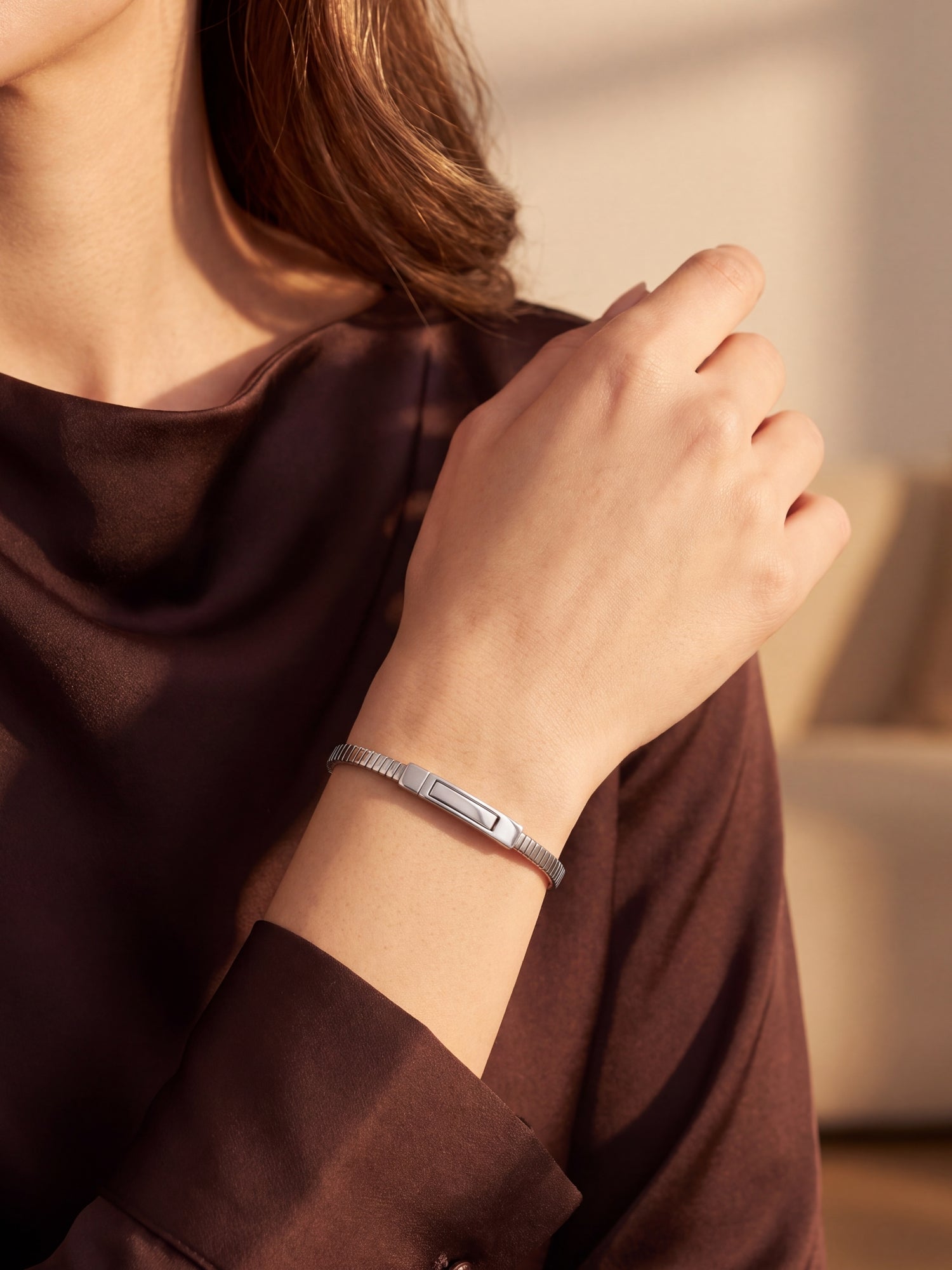 A close-up of a woman wearing a slim silver metal bracelet with a rectangular center detail, paired with a dark brown satin top in warm, sunlit indoor lighting.