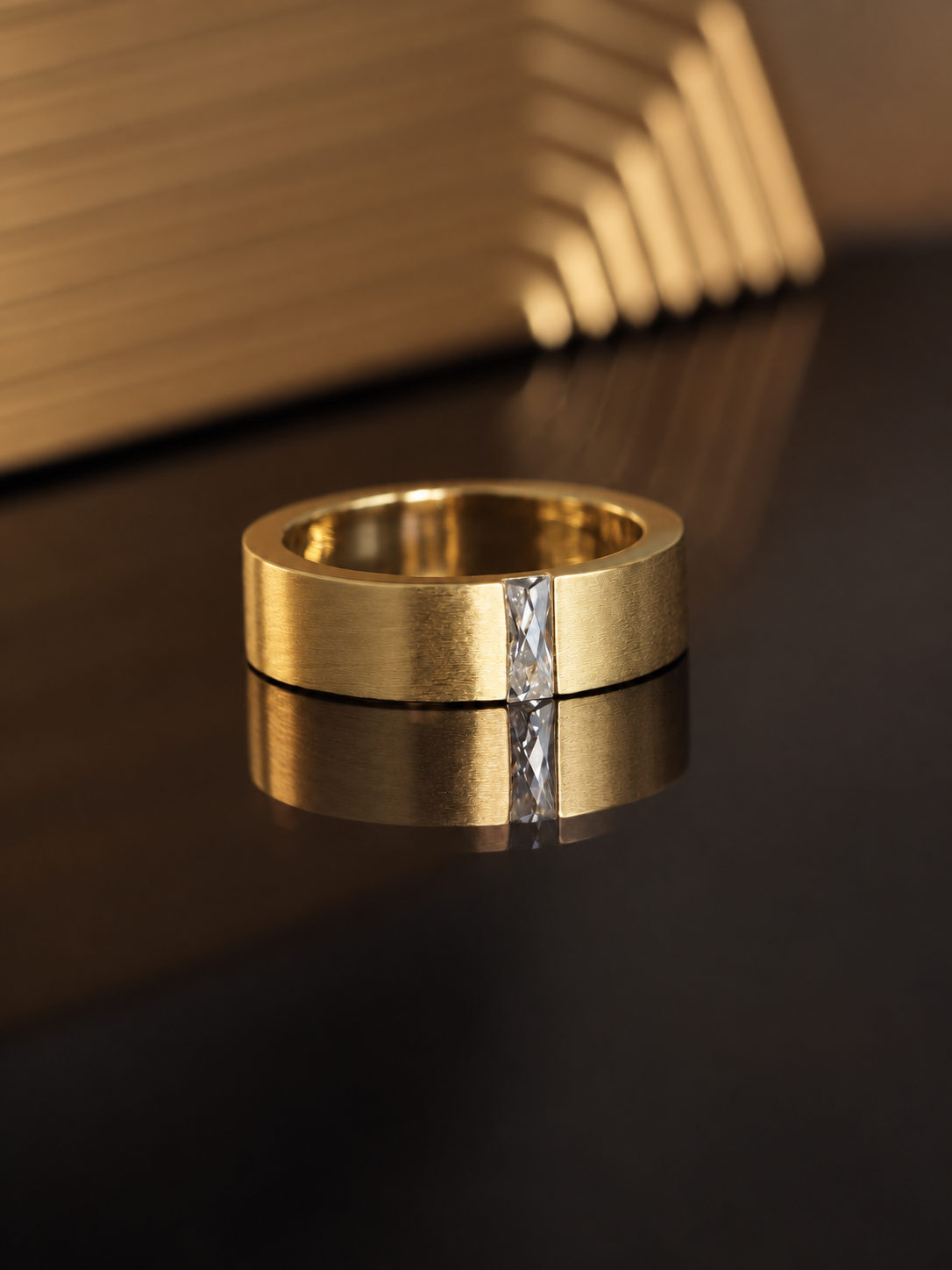 A gold band ring with a single marquise-cut clear gemstone set vertically down the center, resting on a dark reflective surface with a blurred gold geometric background.