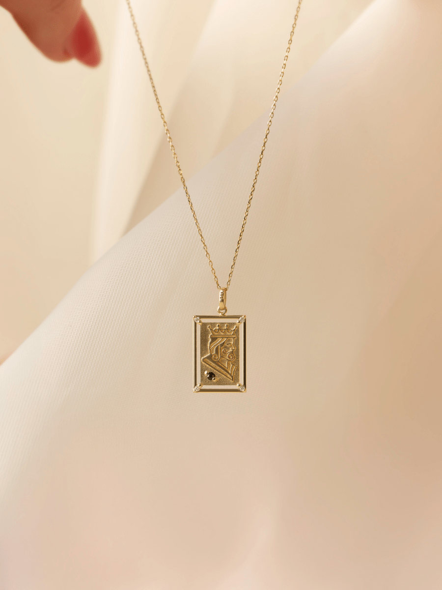 Rectangular playing card charm with embossed gold king illustration and birthstone accent on chain in front of white fabric