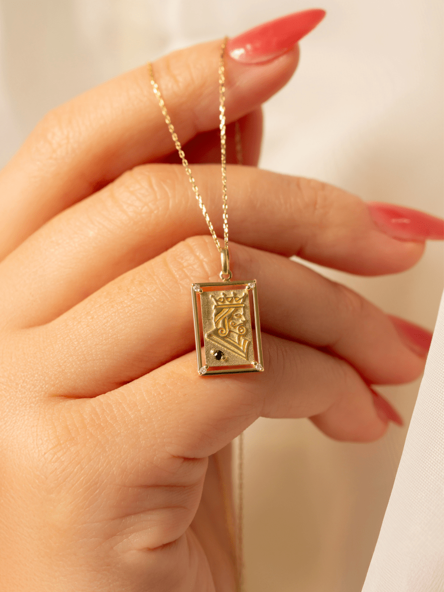 Hand holding rectangular playing card charm with embossed gold king illustration and birthstone accent on chain