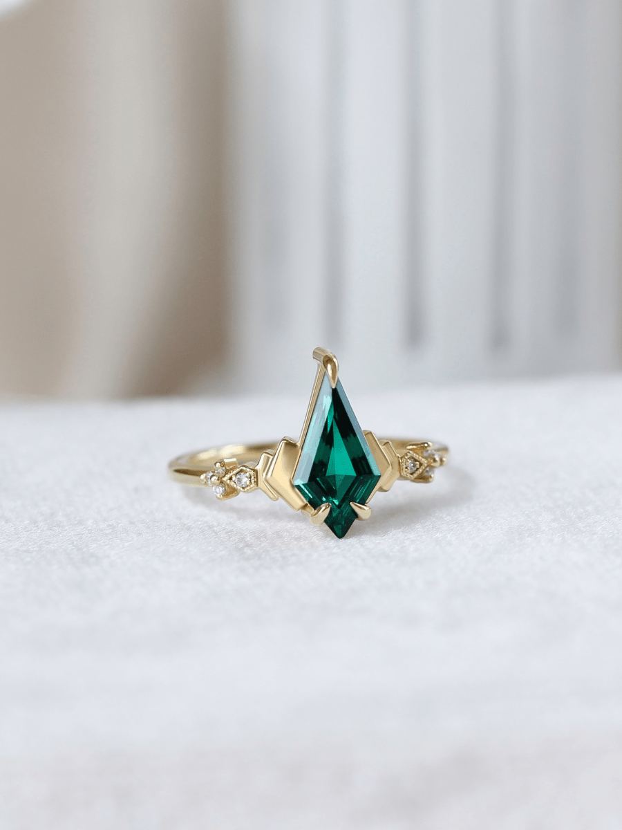 Face of Art Deco kite emerald ring with chevron band in cool ambient light, set against soft white columns evoking an ethereal, timeless feel