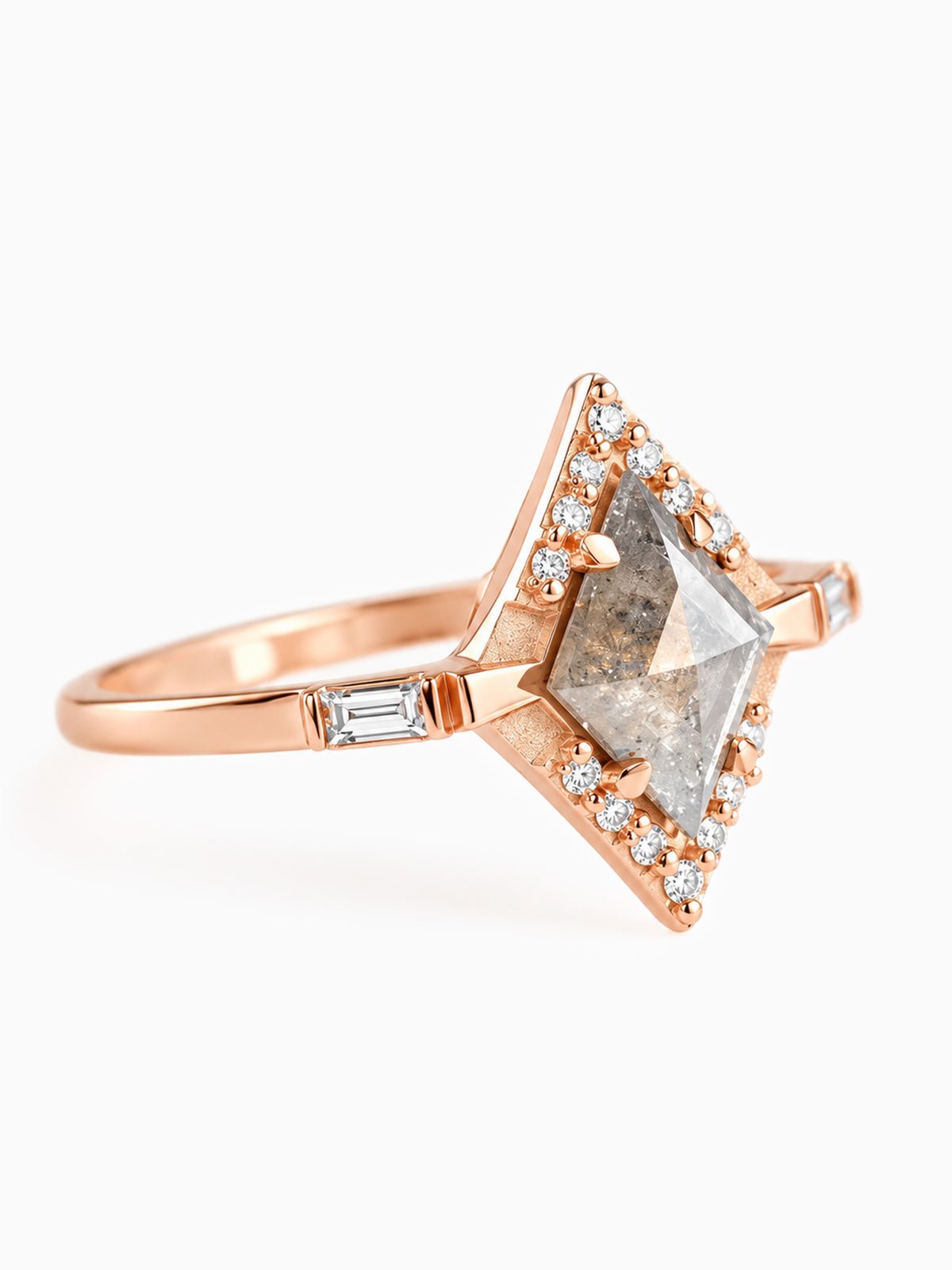 A rose gold ring featuring a large, faceted diamond-cut gray gemstone surrounded by a halo of small clear stones, with a baguette-cut side stone on a clean white background.