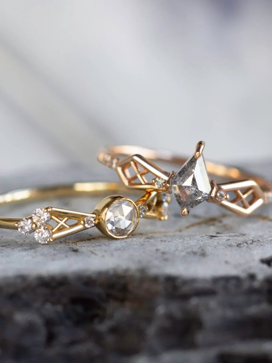 Two modern Art Deco rings with flared bow-shaped geometric shanks and lattice detailing, one with white diamond on yellow gold, one with salt and pepper galaxy diamond on rose gold band, displayed on grey stone