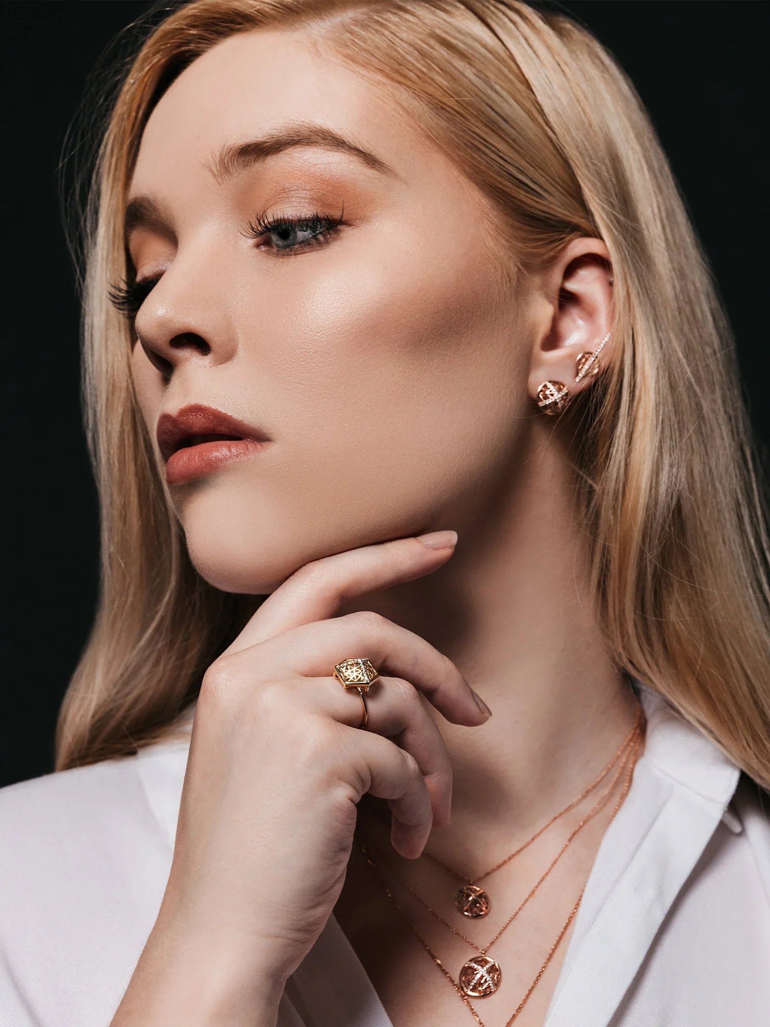 Woman wearing gold jewelry including earrings, ring, and necklace against a black background