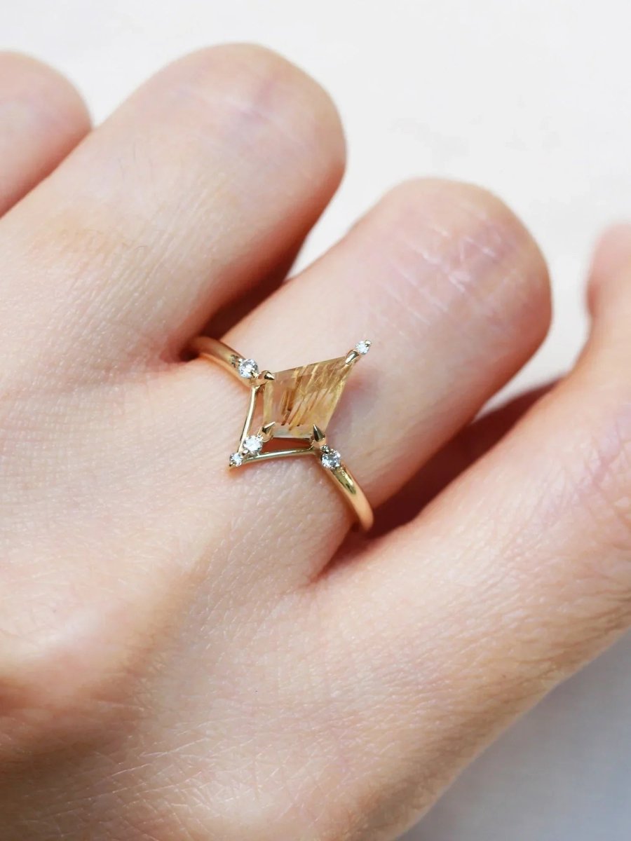 Kite shape rutilated quartz engagement ring with smaller round diamonds in 14k gold inspired by the art deco style and minimalism on model's hand.