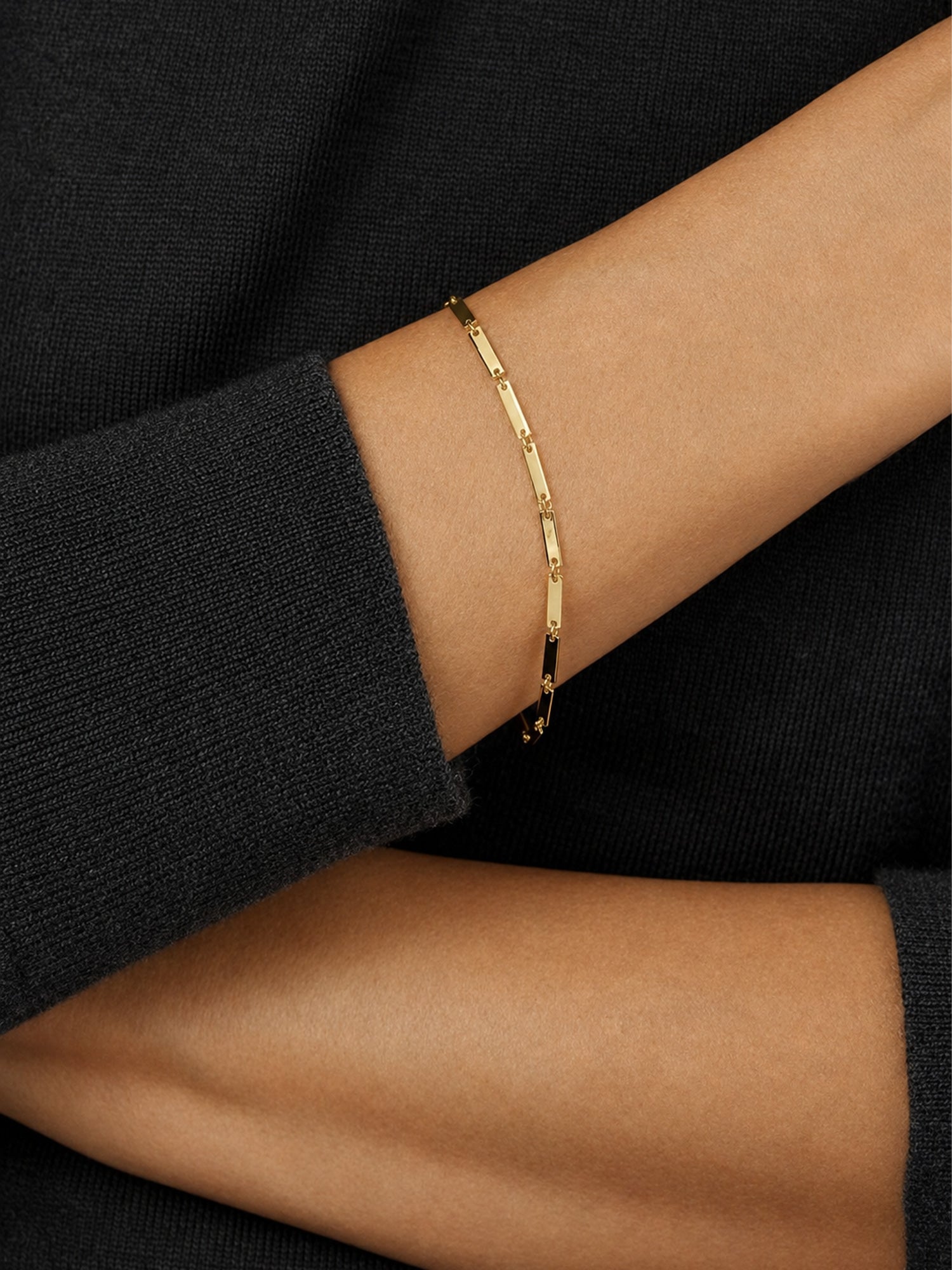A delicate gold chain bracelet with small, evenly spaced oval links, worn on a person's wrist against a black knit sweater.