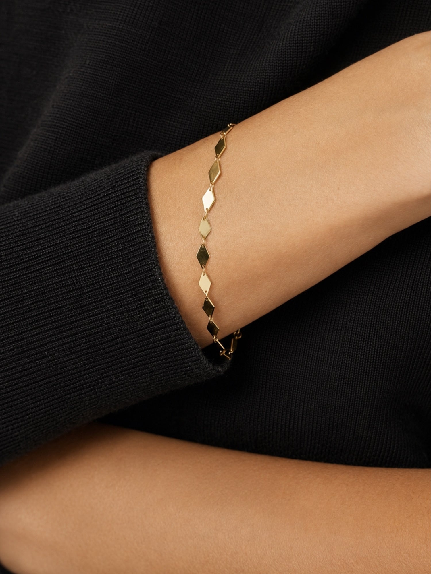 A delicate gold chain bracelet with faceted Kite diamond-shaped links, shown worn on a person's wrist against a black knit sweater.