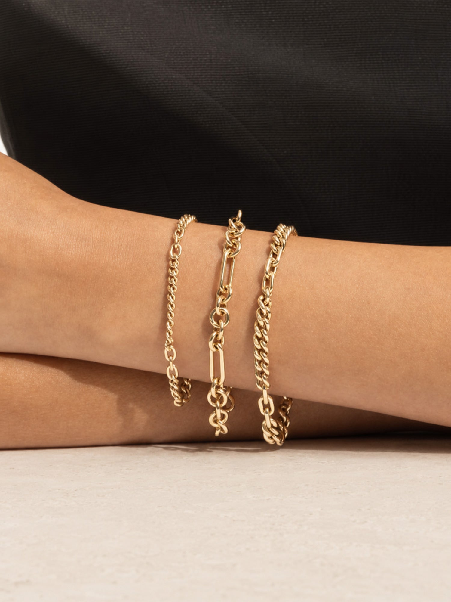 Three gold chain bracelets with different link styles worn on a person's wrist, shown against a black top and light surface.