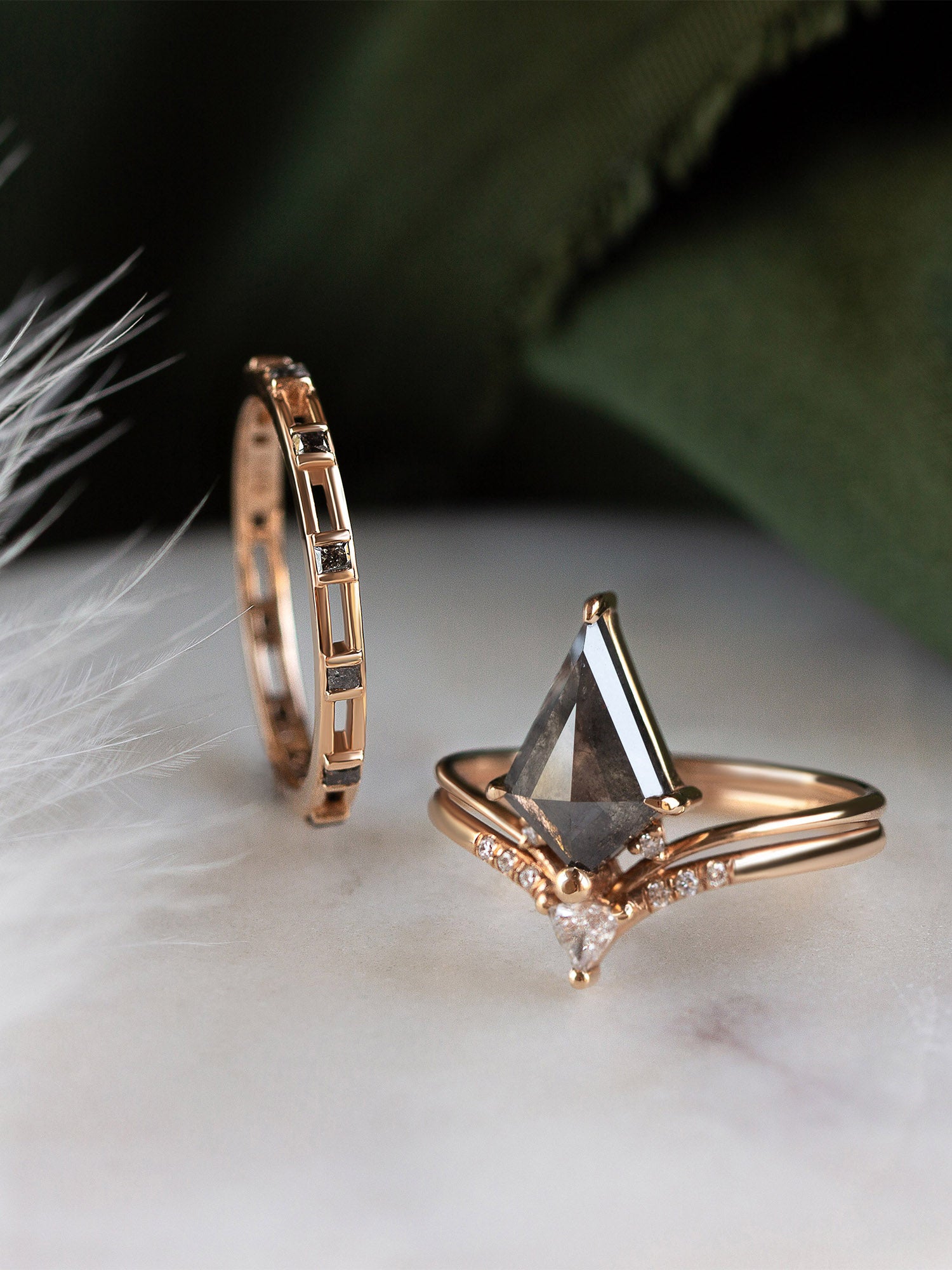 A pair of rose gold rings displayed on a white marble surface with soft white feathers and a dark green fabric background. One ring features a large triangular clear gemstone with a pointed diamond accent, while the other is a slim geometric band with small dark inset stones.