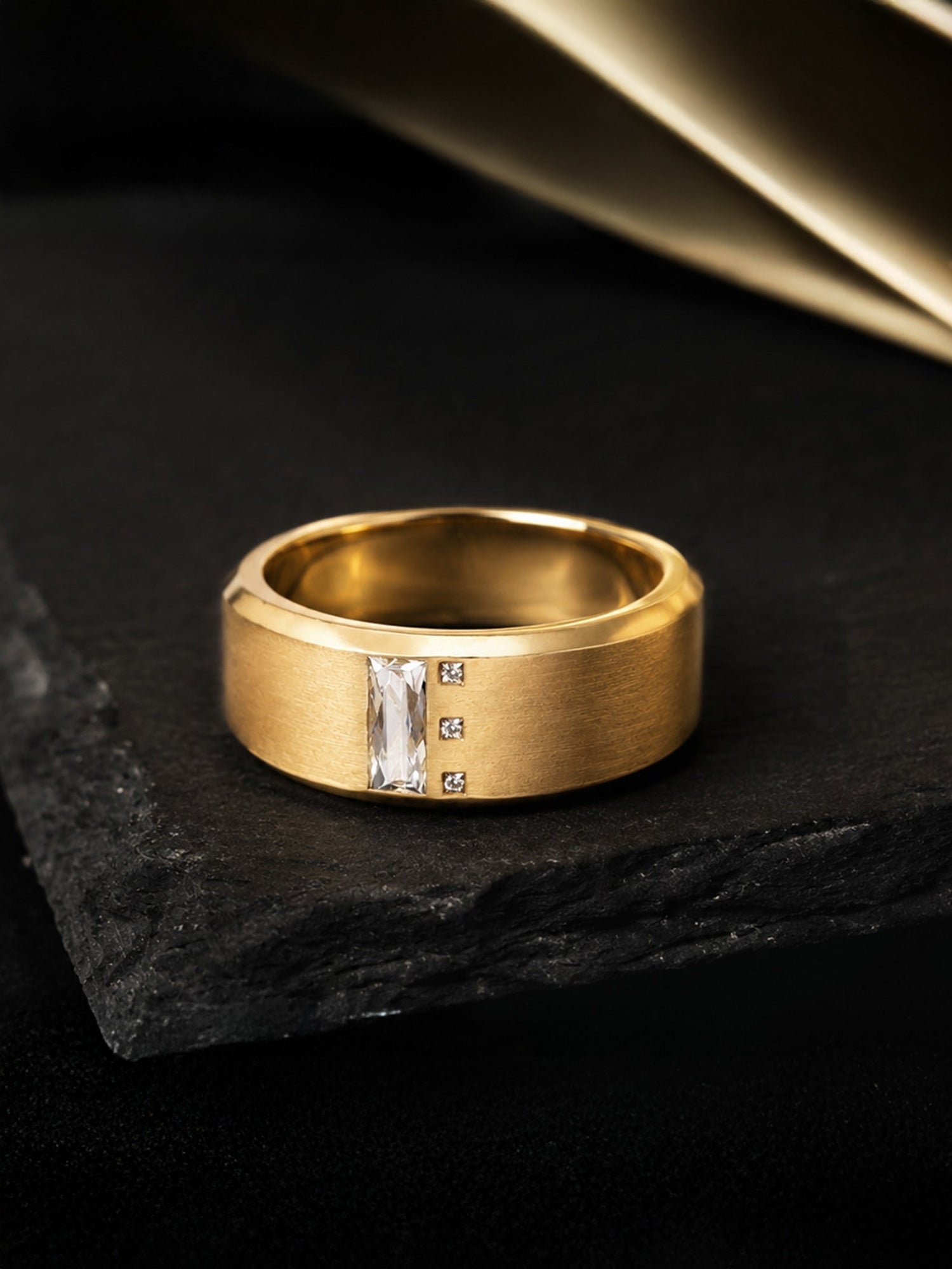A gold band ring with a central vertical baguette-cut clear gemstone and three small round clear stones, displayed on a dark textured stone surface.
