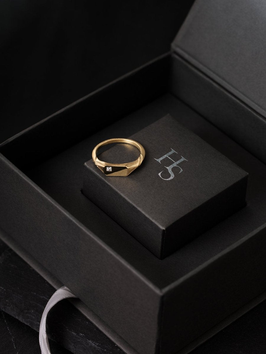 Gold ring with a diamond on a black box with "HS" initials, set against a dark background.