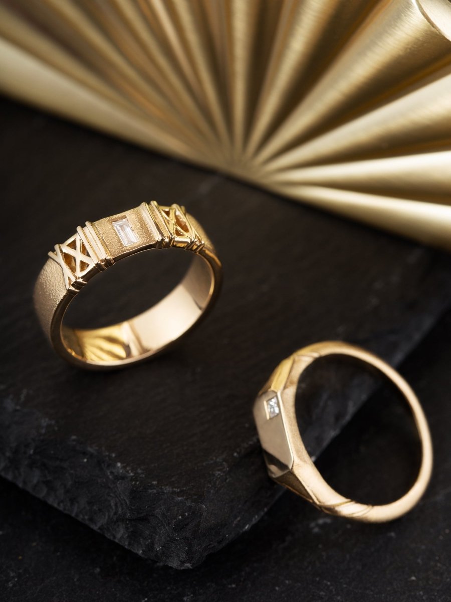 Two gold rings with diamond accents on a dark surface with a gold fan in the background.