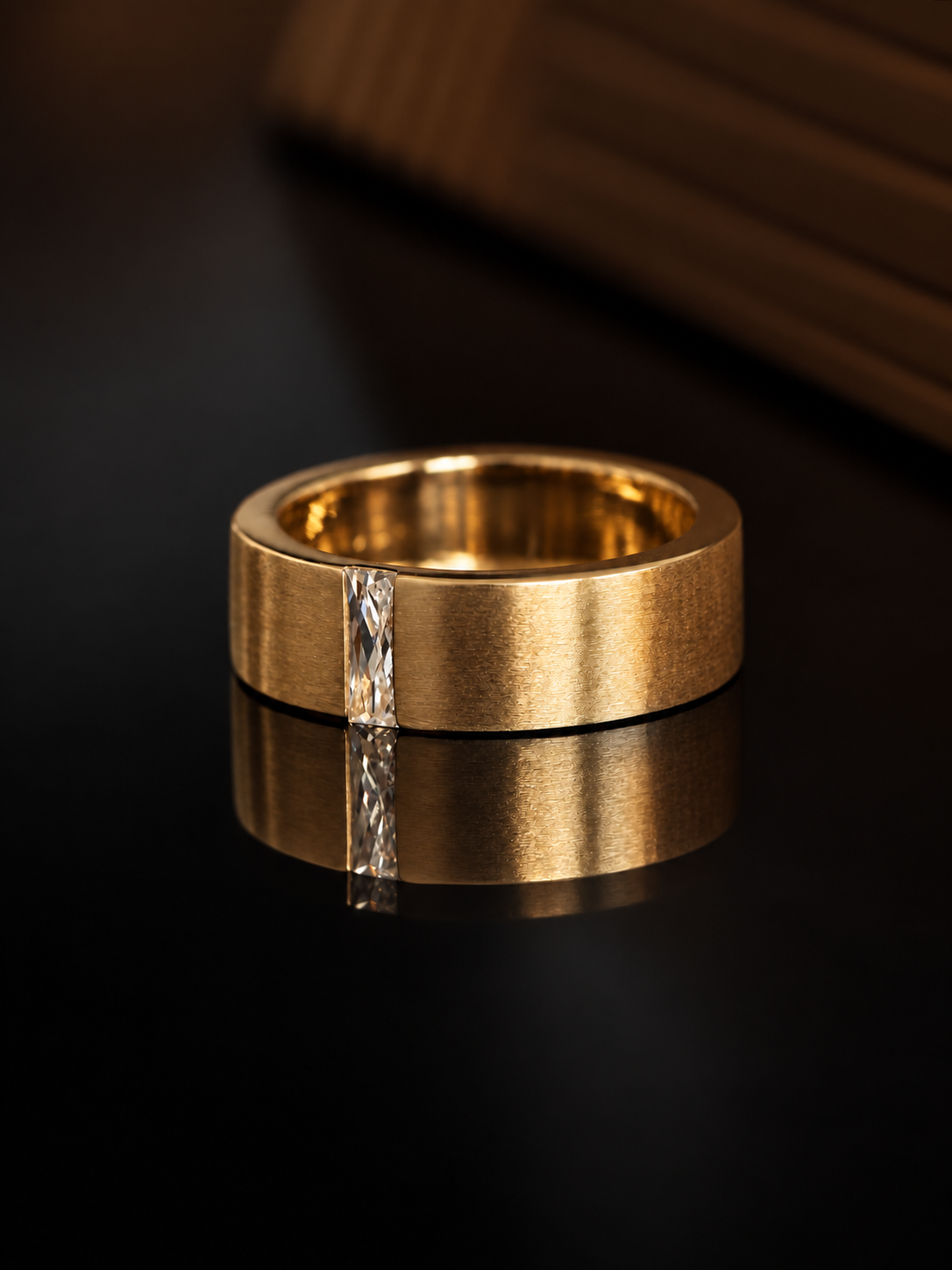 A wide, chunky gold band ring with a single clear baguette-cut gemstone set vertically down the center, resting on a glossy black reflective surface.