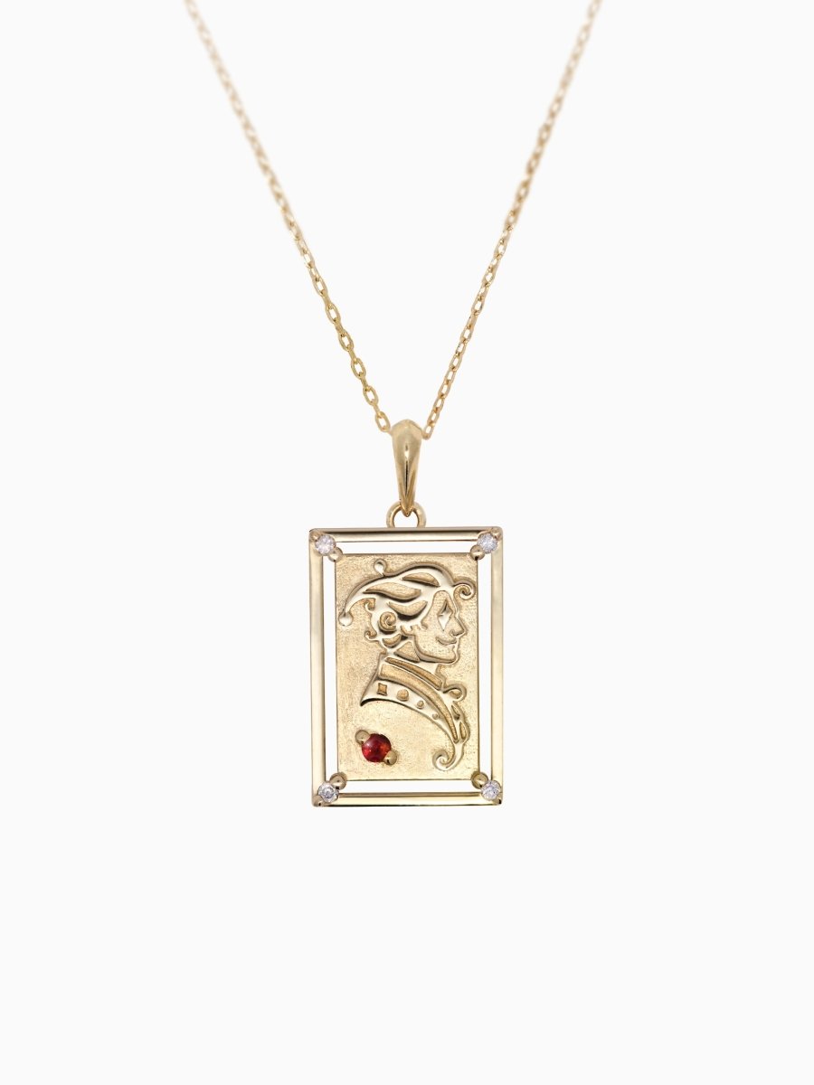A gold necklace featuring a rectangular charm with an engraved design resembling a playing card's joker, with a red gemstone at the bottom.