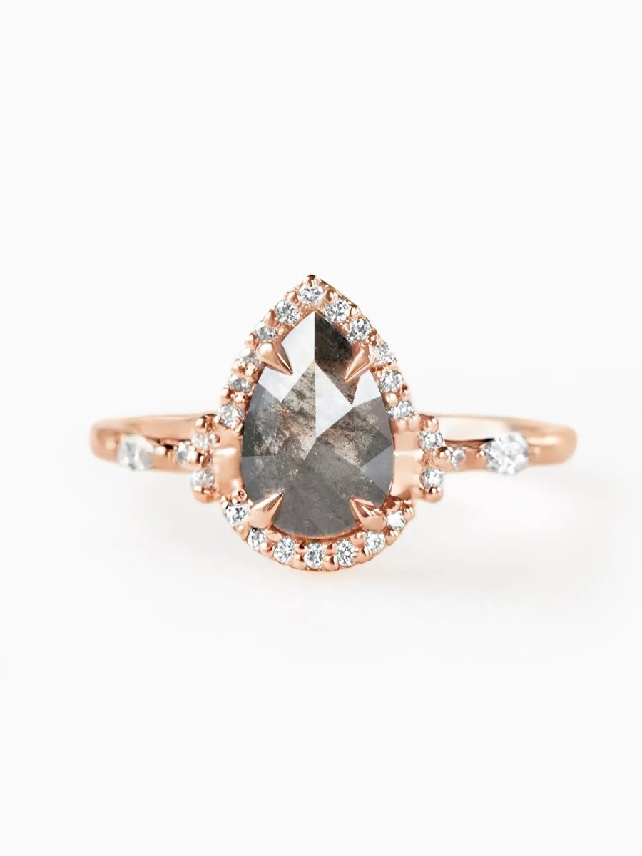 A pear-shaped diamond ring with a salt and pepper appearance, set in a rose gold band and surrounded by smaller clear diamonds creating a halo effect.