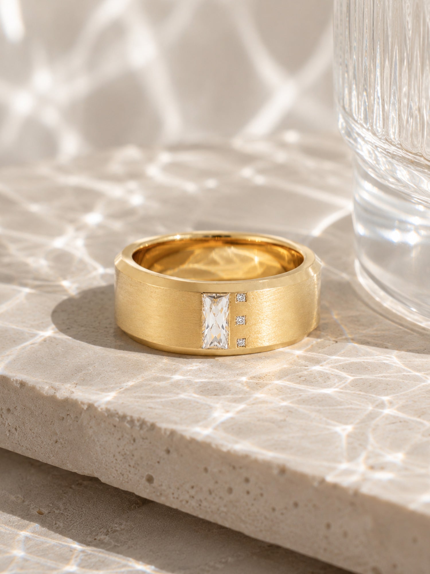 A wide yellow gold ring with a rectangular baguette-cut clear center stone and two small side stones, displayed on a textured stone surface with dappled light.