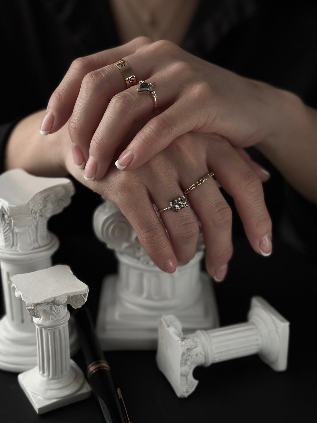 A close-up of a person's hands resting on a dark surface, adorned with multiple delicate rings. The hands are styled with white French manicure nails, and the background features decorative white classical column fragments against a dark backdrop.